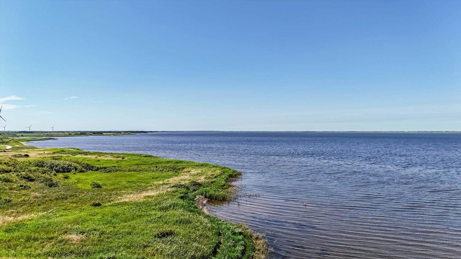 Beach with sand, stones, and grass by the water. Wind turbines are visible in the background.