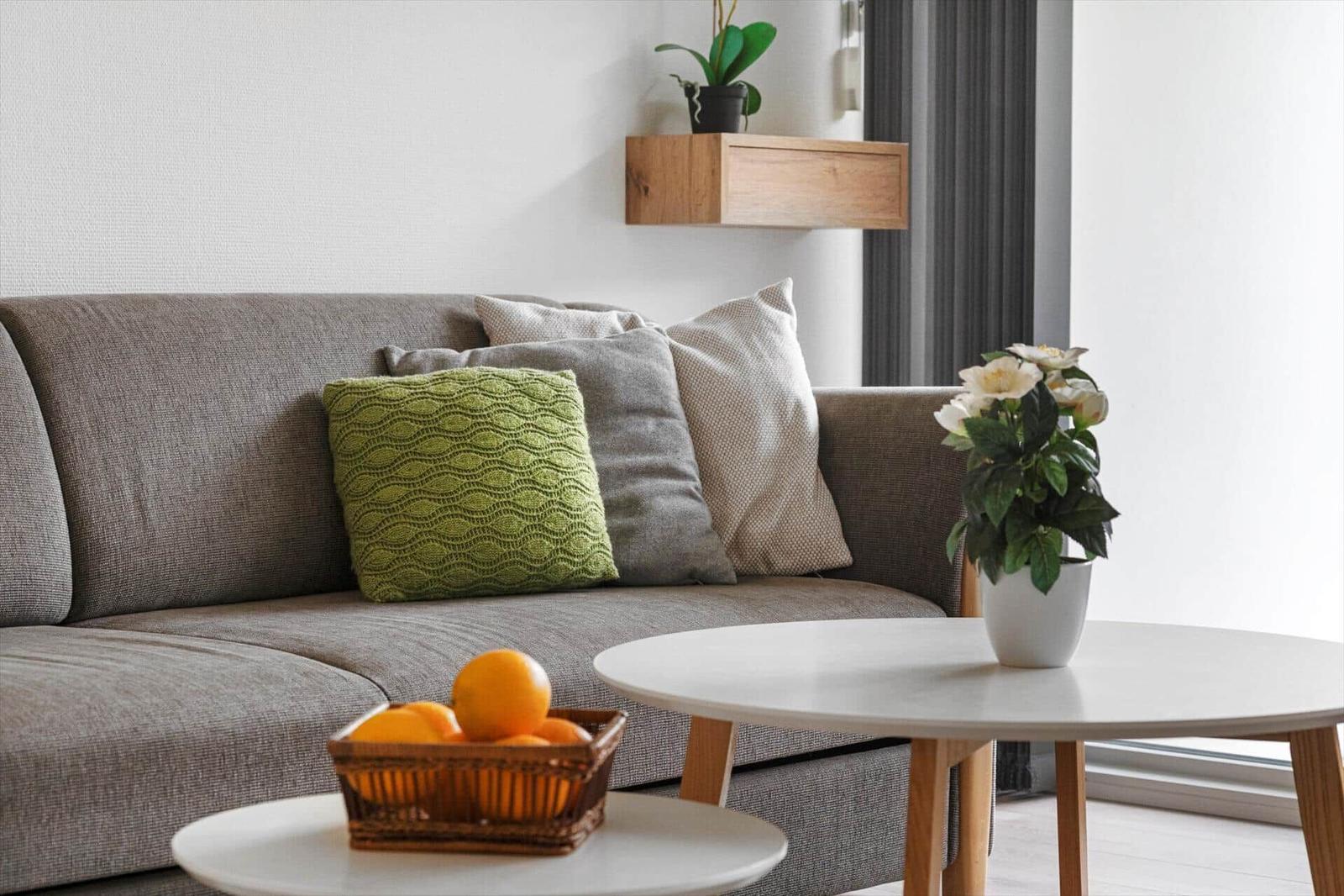 Grauer Sofa mit Kissen, runder Tisch, Obstkorb und Pflanzen.