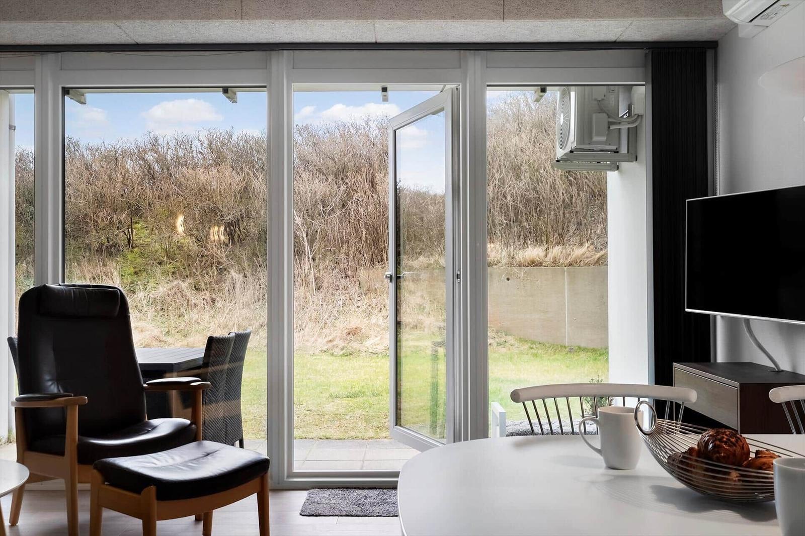 Ein Raum mit großem Fensterblick auf Grünflächen und Bäume. Es gibt einen Sessel, einen Tisch und eine TV-Anlage.