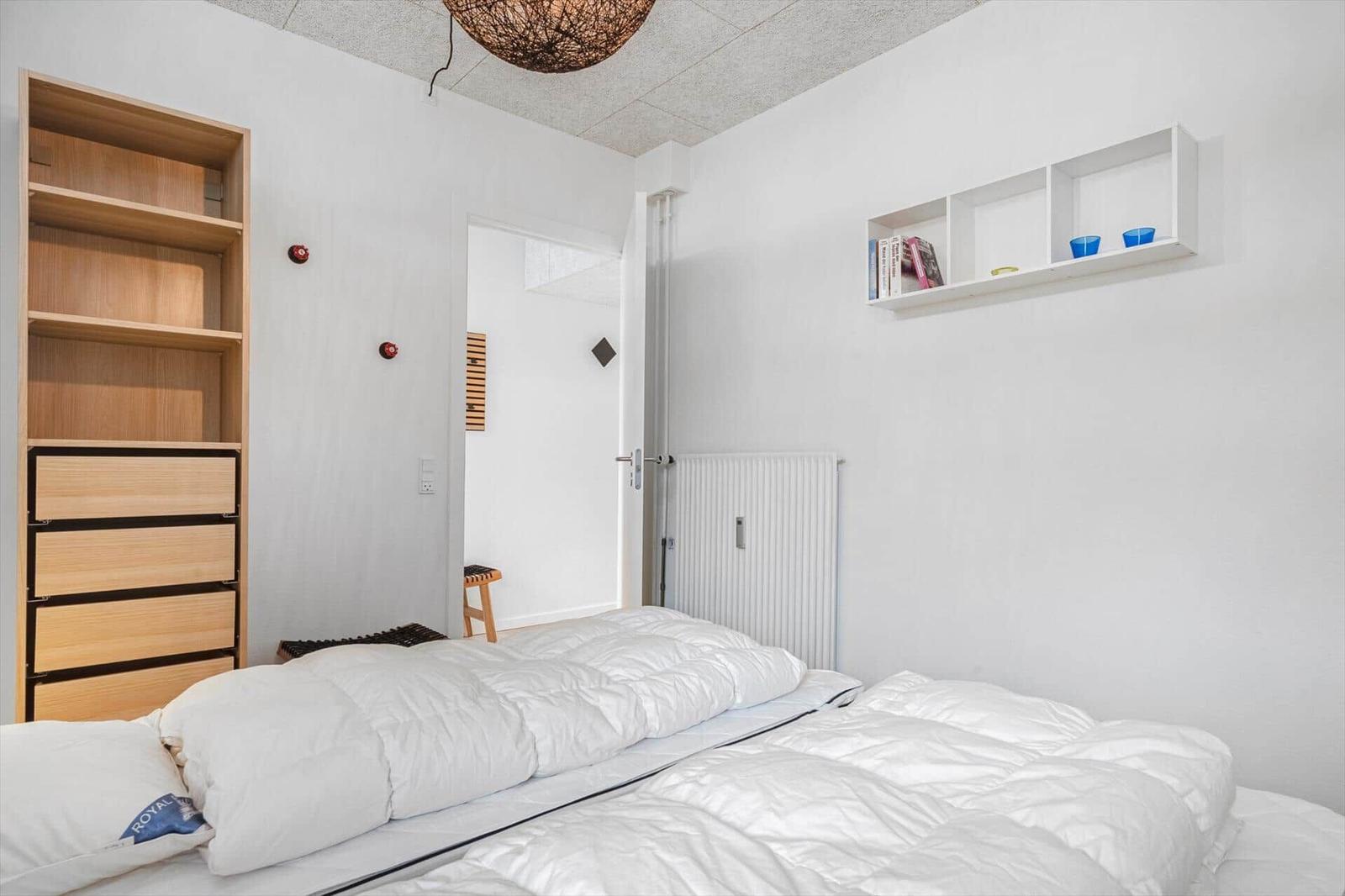 Schlafzimmer mit weißem Bett, Holzregal und Wandregal mit Büchern und Tassen.