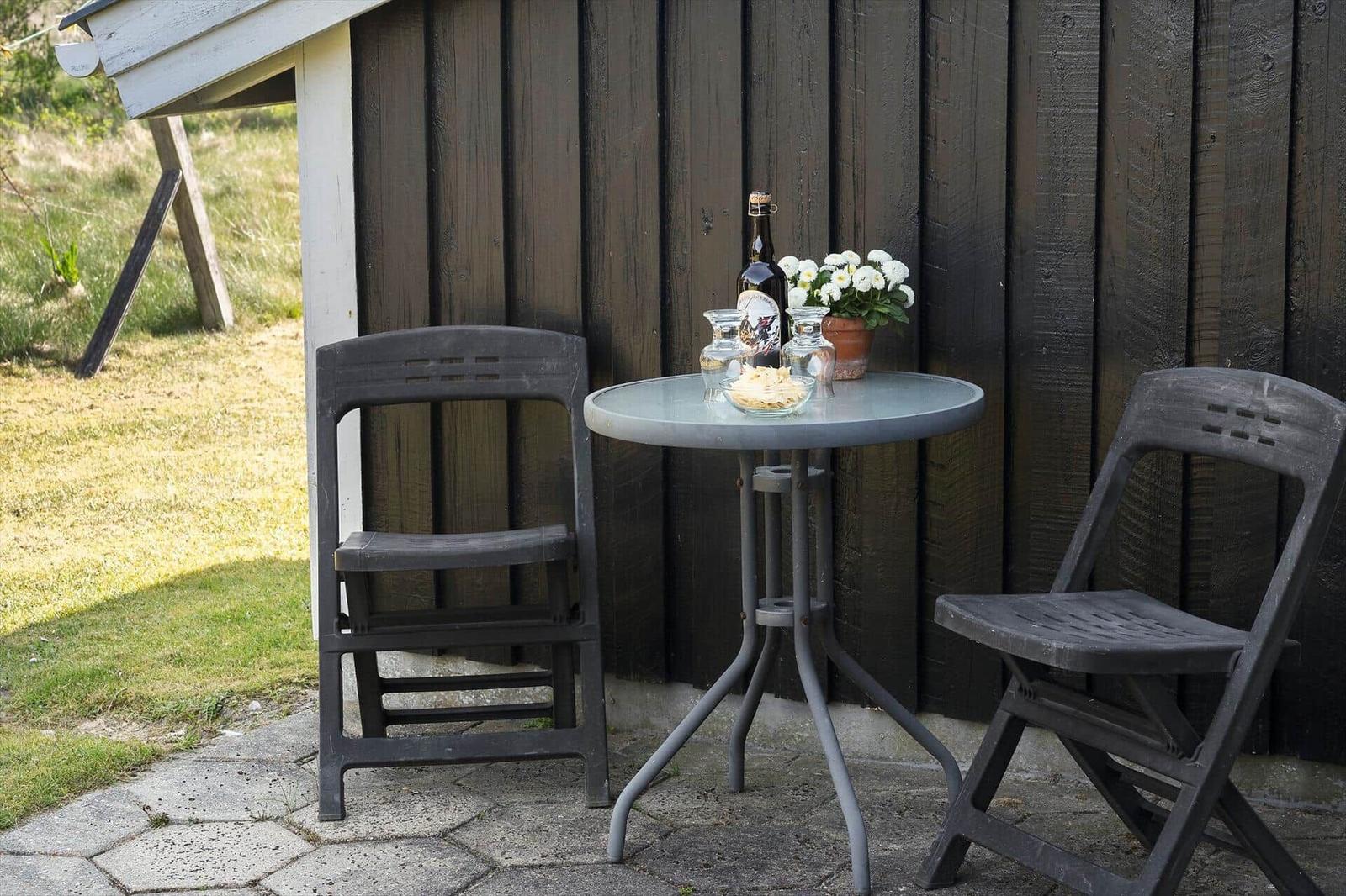 Rundtisch mit zwei Stühlen, Flasche, Gläser und Blumen vor Holzwand.