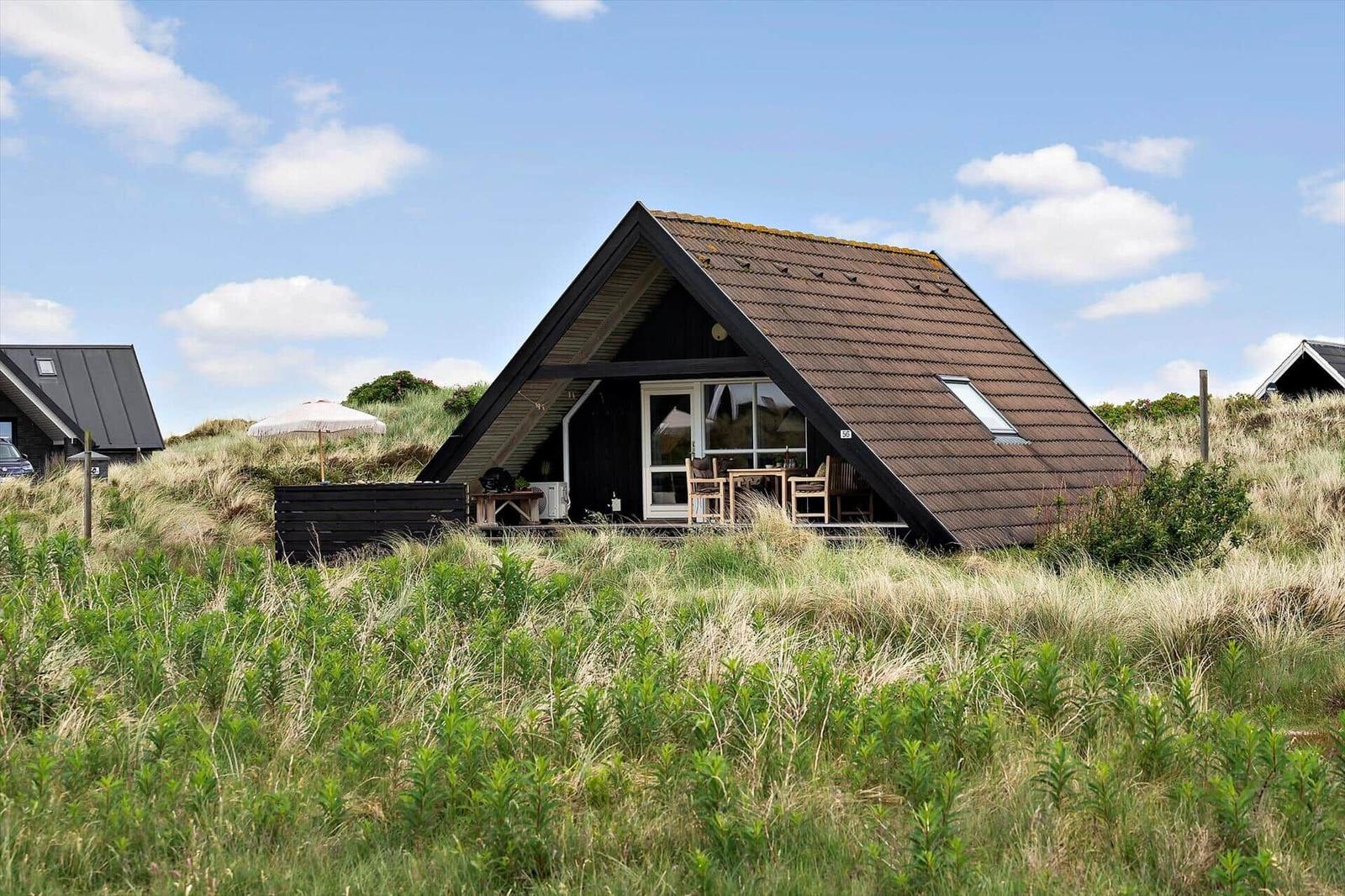 A-frame-Haus mit Terrasse, Gartenmöbeln und Sonnenschirm in Graslandschaft.
