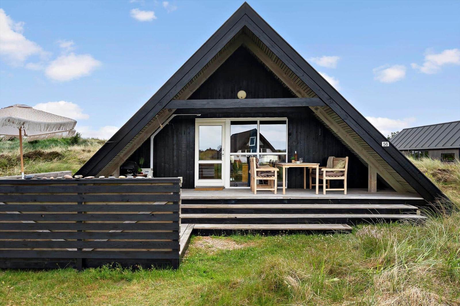 Schwarze Holzhütte mit Terrasse, Tisch und Stühlen. Hintergrund: Gras und Himmel.