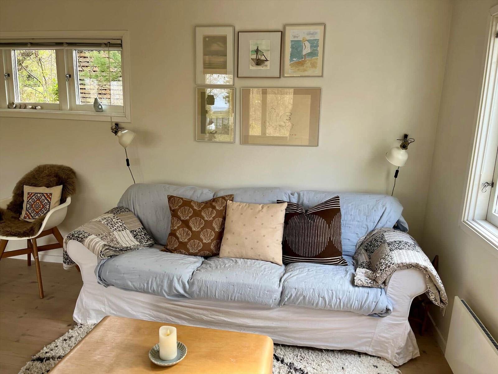 Ein Wohnzimmer mit Sofa, Kissen, Tisch und Fenstern. An der Wand hängen Bilder.