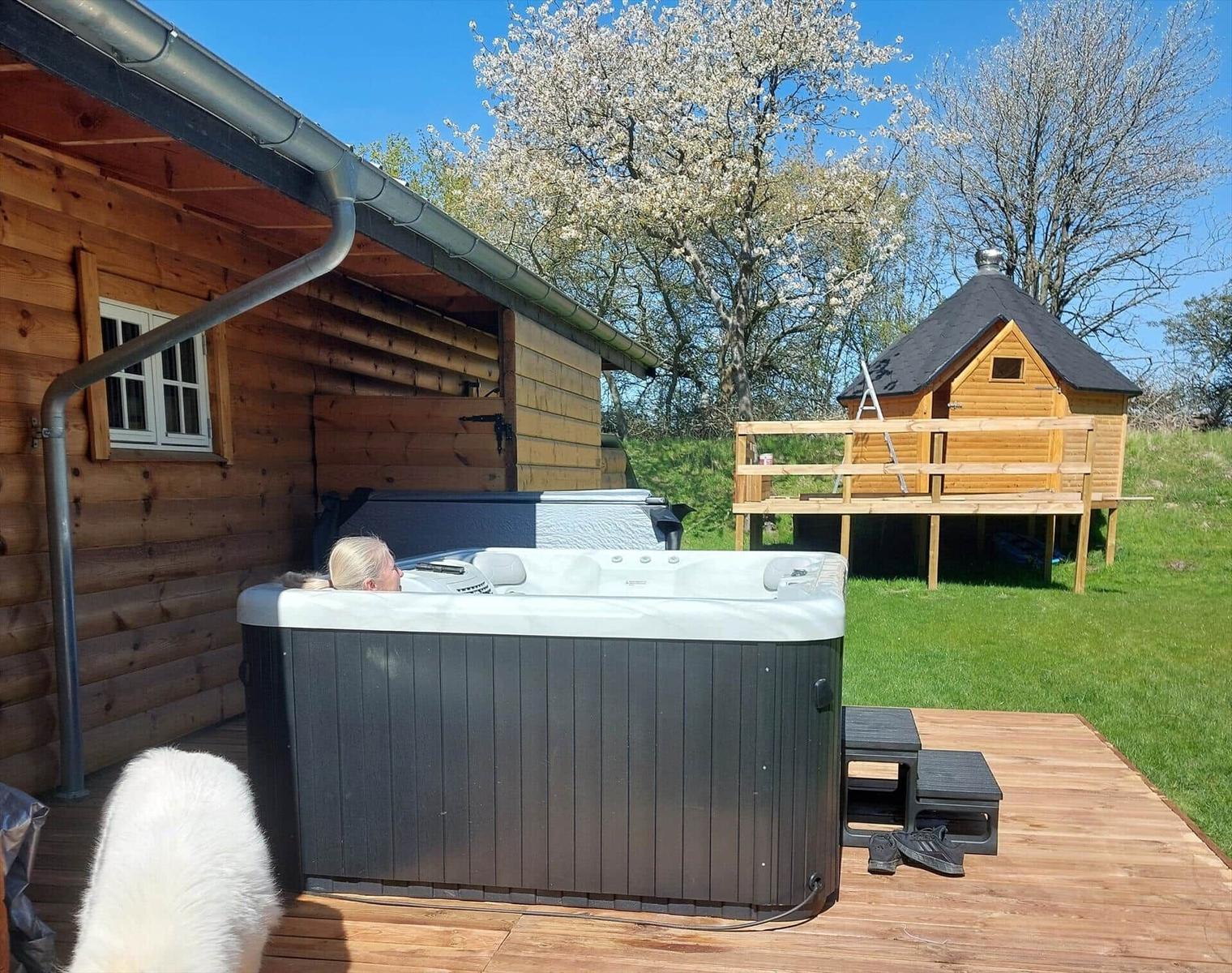 A person relaxes in a hot tub on a deck next to a wooden house. Another wooden structure is visible in the background.