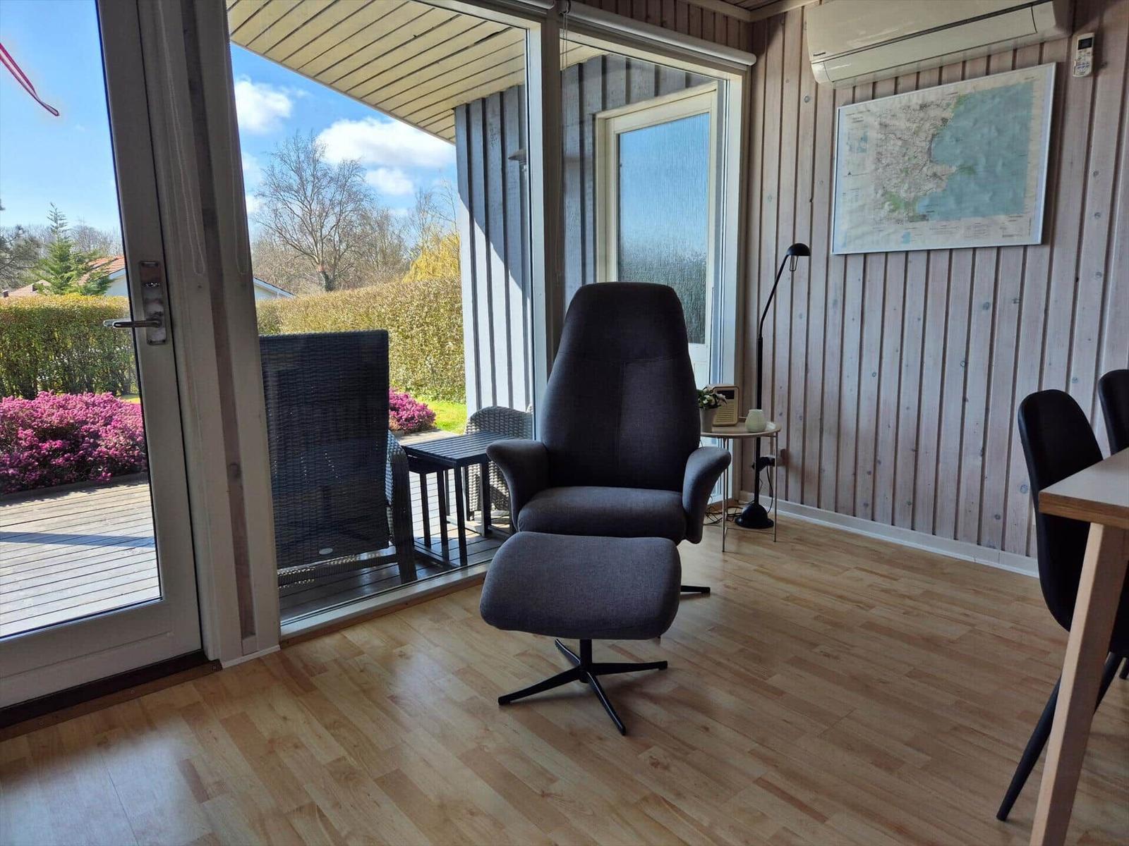 Ein grauer Sessel mit Fußstütze steht im Raum. Durch die Glastür ist eine Terrasse mit Garten zu sehen.