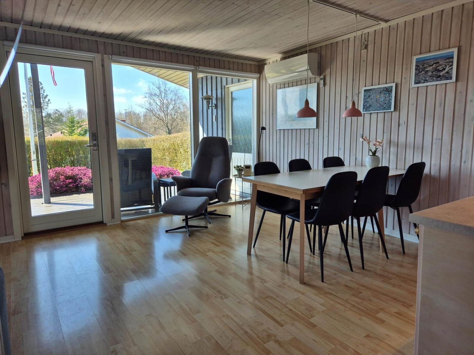 Essbereich mit Tisch, Stühlen und Sessel. Holzfußboden und Holzwandverkleidung. Glastüren zum Außenbereich.
