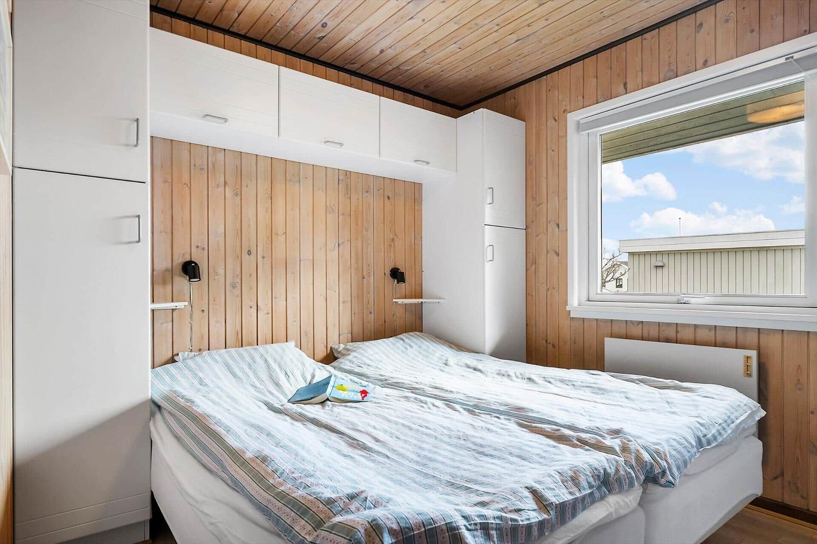 Bedroom with double bed, wooden wall, and window. White cabinets and nightstands.