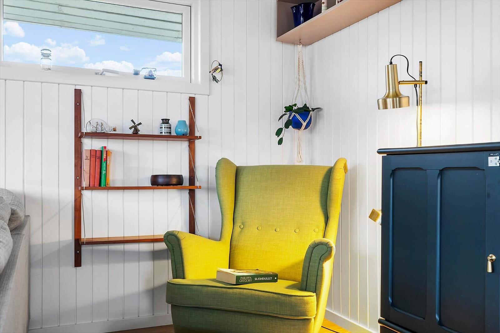 A yellow armchair is positioned by a window with white walls and a wooden shelf.