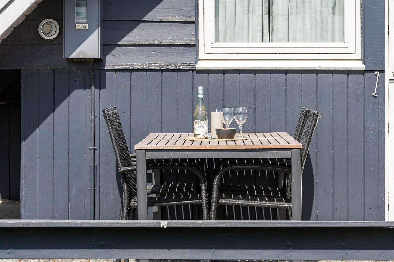Außenbereich mit Tisch und Stühlen vor grauem Holzhaus. Auf Tisch stehen Weinflasche und Gläser.
