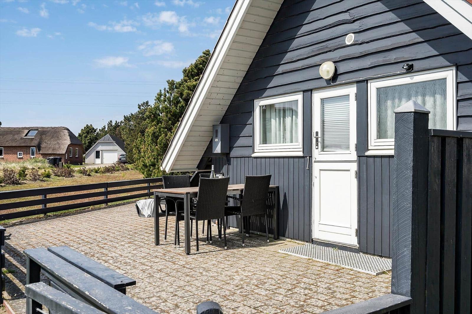Terrasse mit Tisch und Stühlen vor einem Haus mit dunkler Fassade und weißer Tür.
