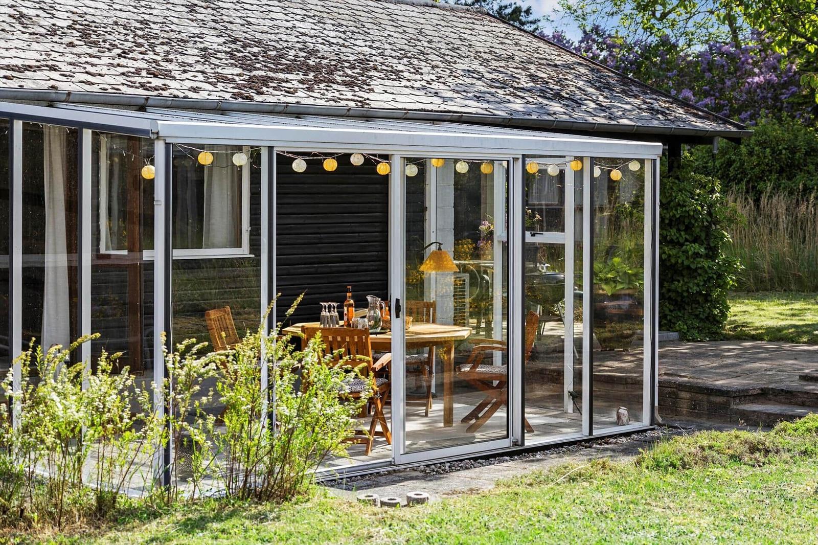 Glass conservatory with wooden table and chairs, string lights, and garden in background.