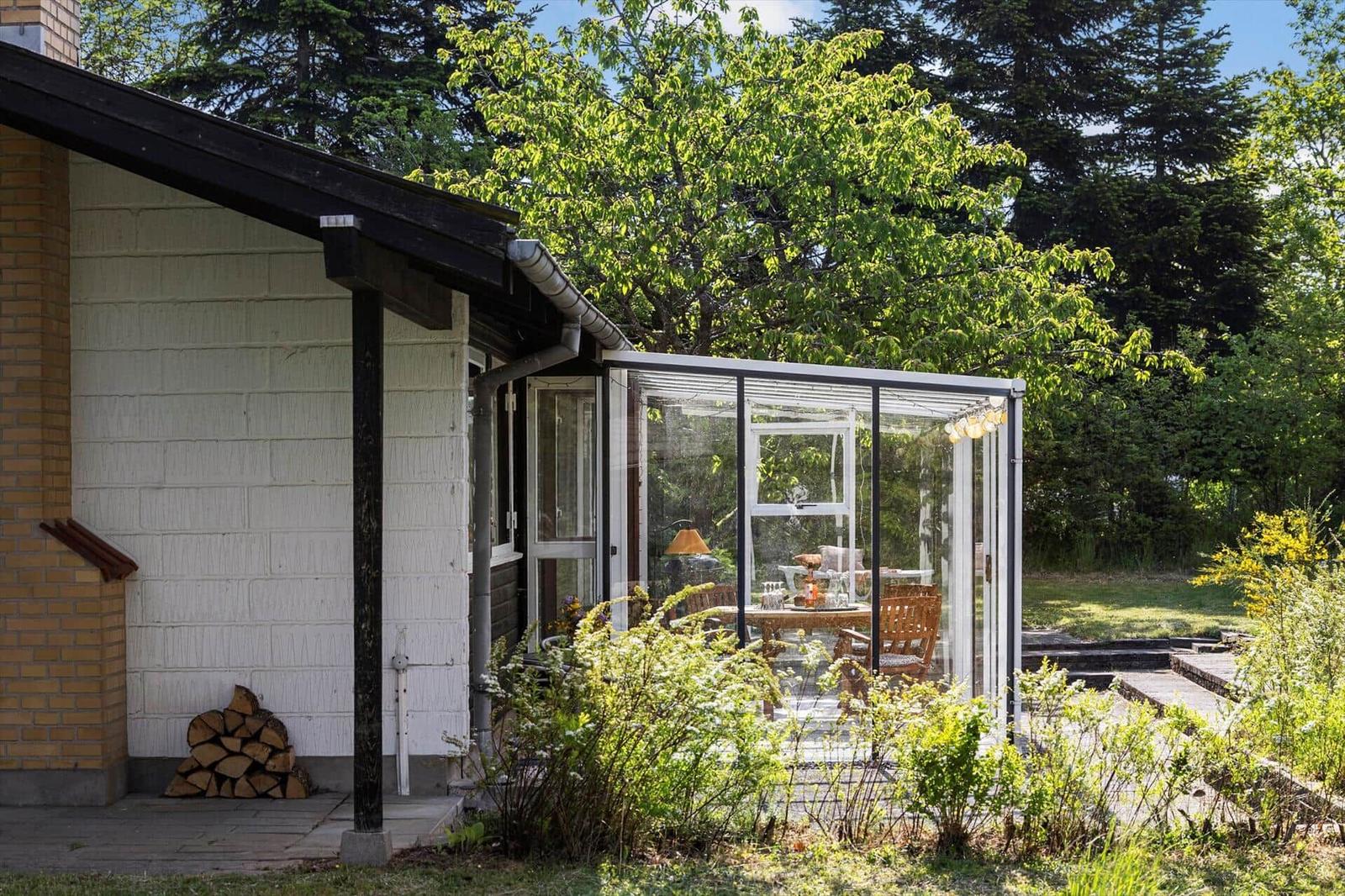 Patio with glass conservatory, wooden table and chairs. Backyard with greenery and trees.