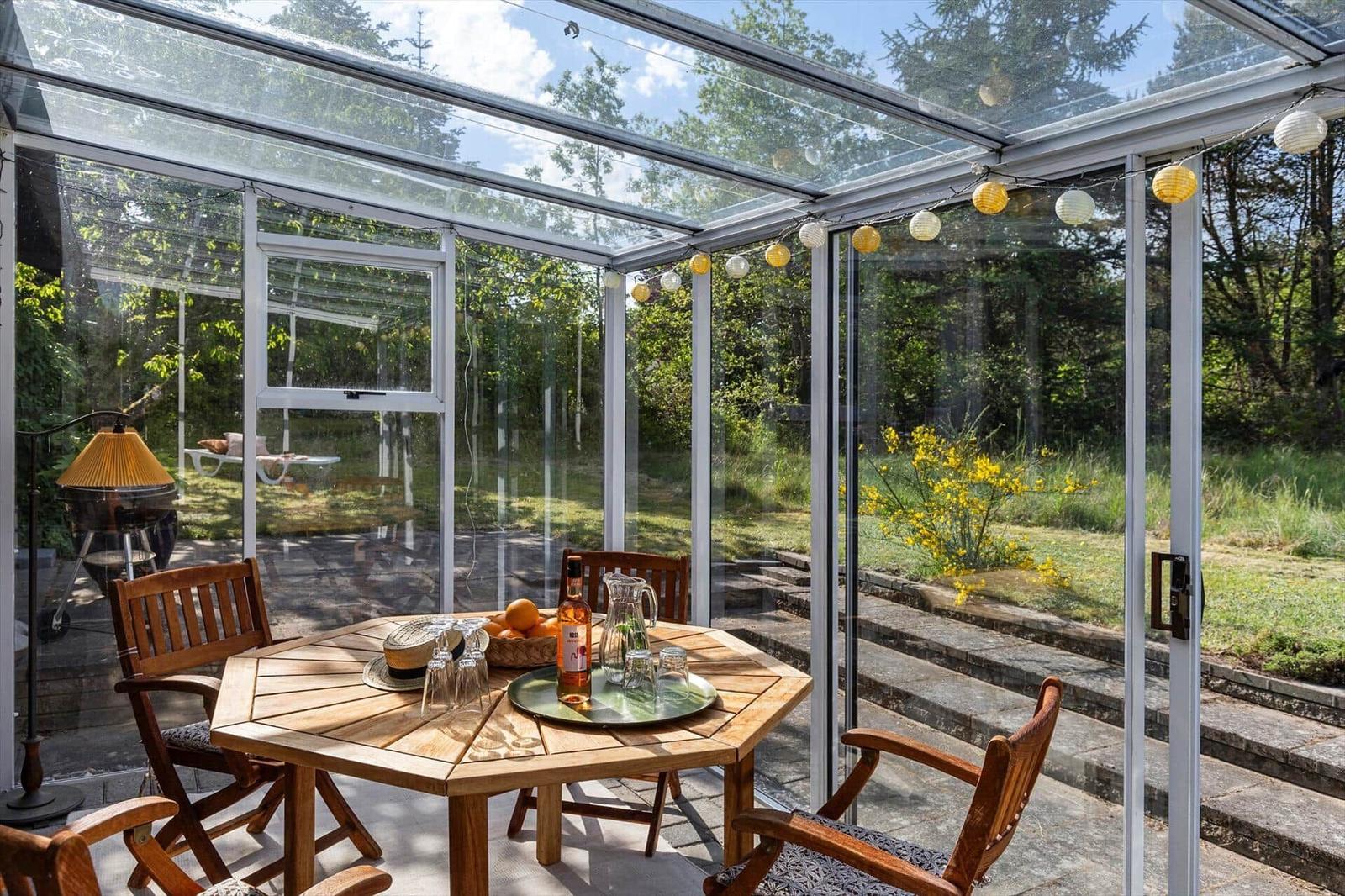 Glass conservatory with wooden table, chairs, and string lights. Outdoor area with greenery and steps.