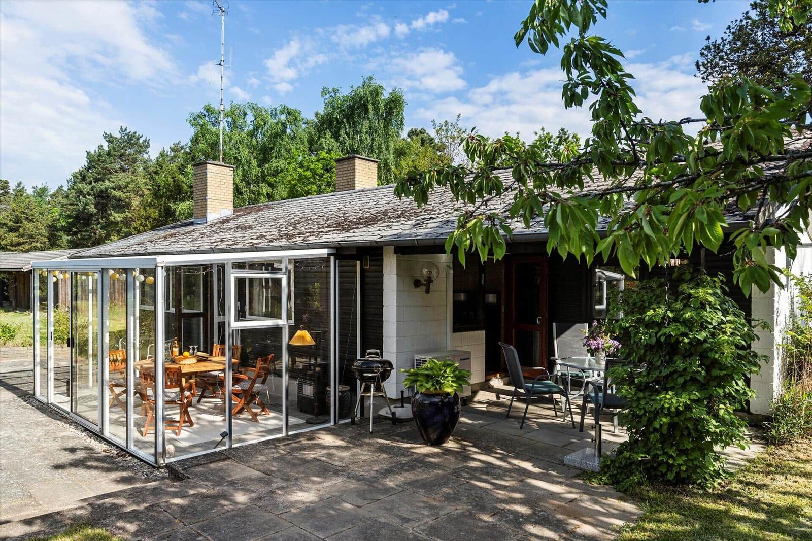 House with glass conservatory, patio, and grill. Surrounded by trees and greenery.