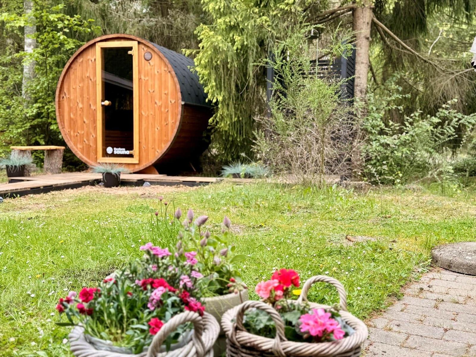 Round wooden sauna with open door in greenery, foreground flower baskets on lawn.