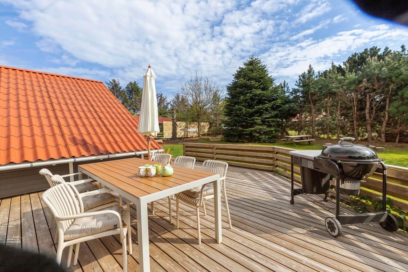 Deck with table, chairs, umbrella, and grill. Background: forest and roof.