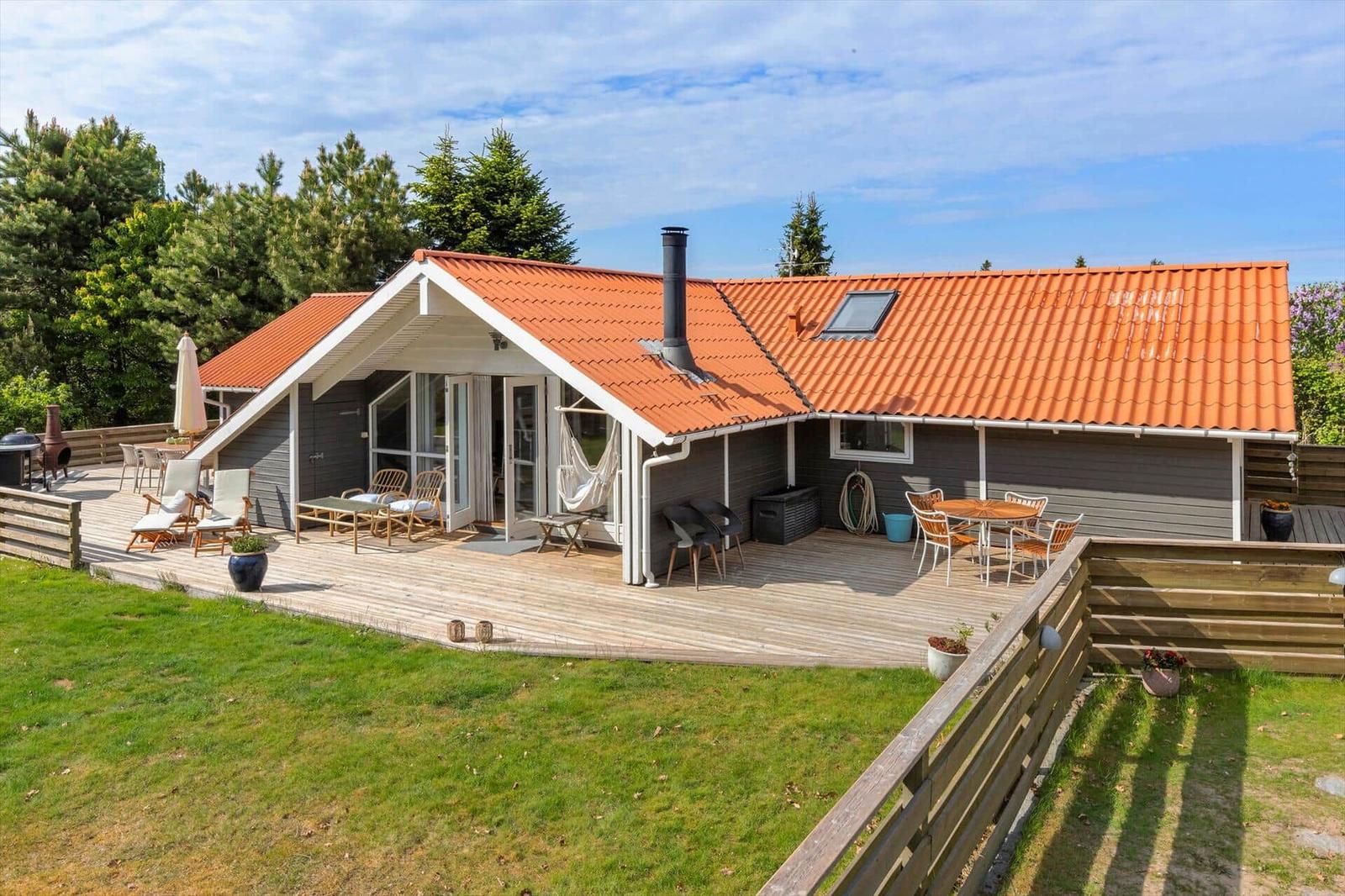 House with wooden terrace, garden, and grill area. Red tiled roof. Terrace with seating.