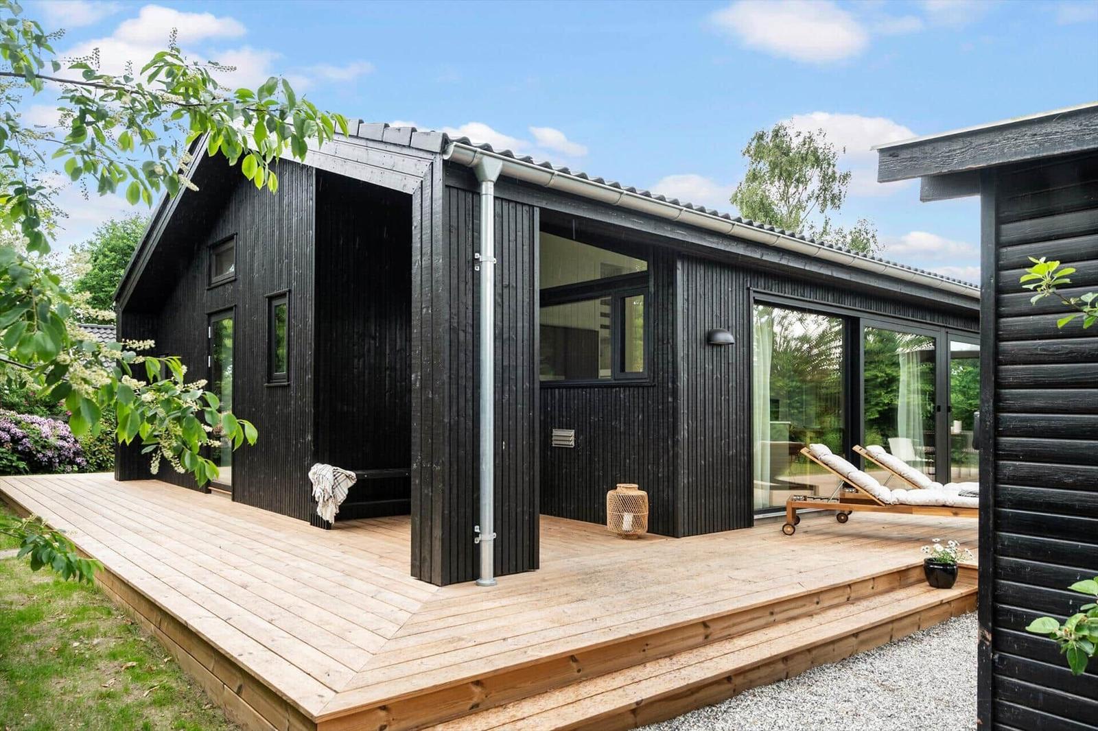 Black wooden house with terrace and loungers. Large windows and covered entrance area.