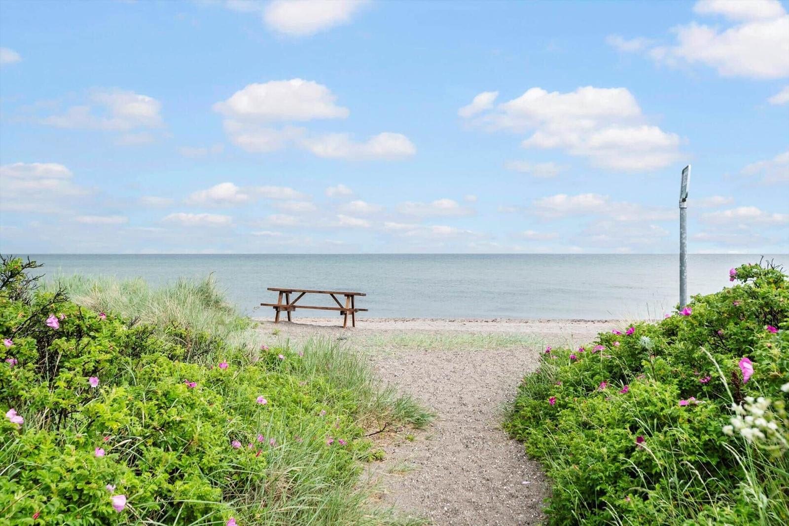 A wooden table stands on the beach, surrounded by grass and pink flowers under a blue sky.