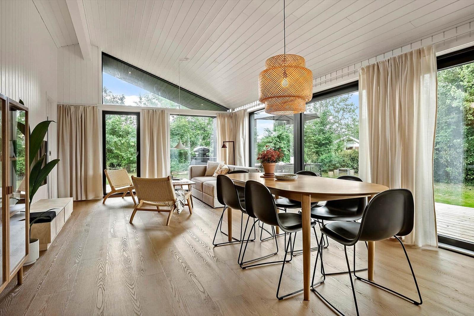 Dining area with wooden table and chairs, adjacent living area with sofa and large windows.