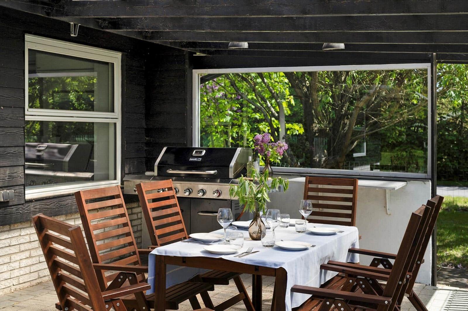 Terrace with table, chairs, grill, and flower arrangement. View of green trees.