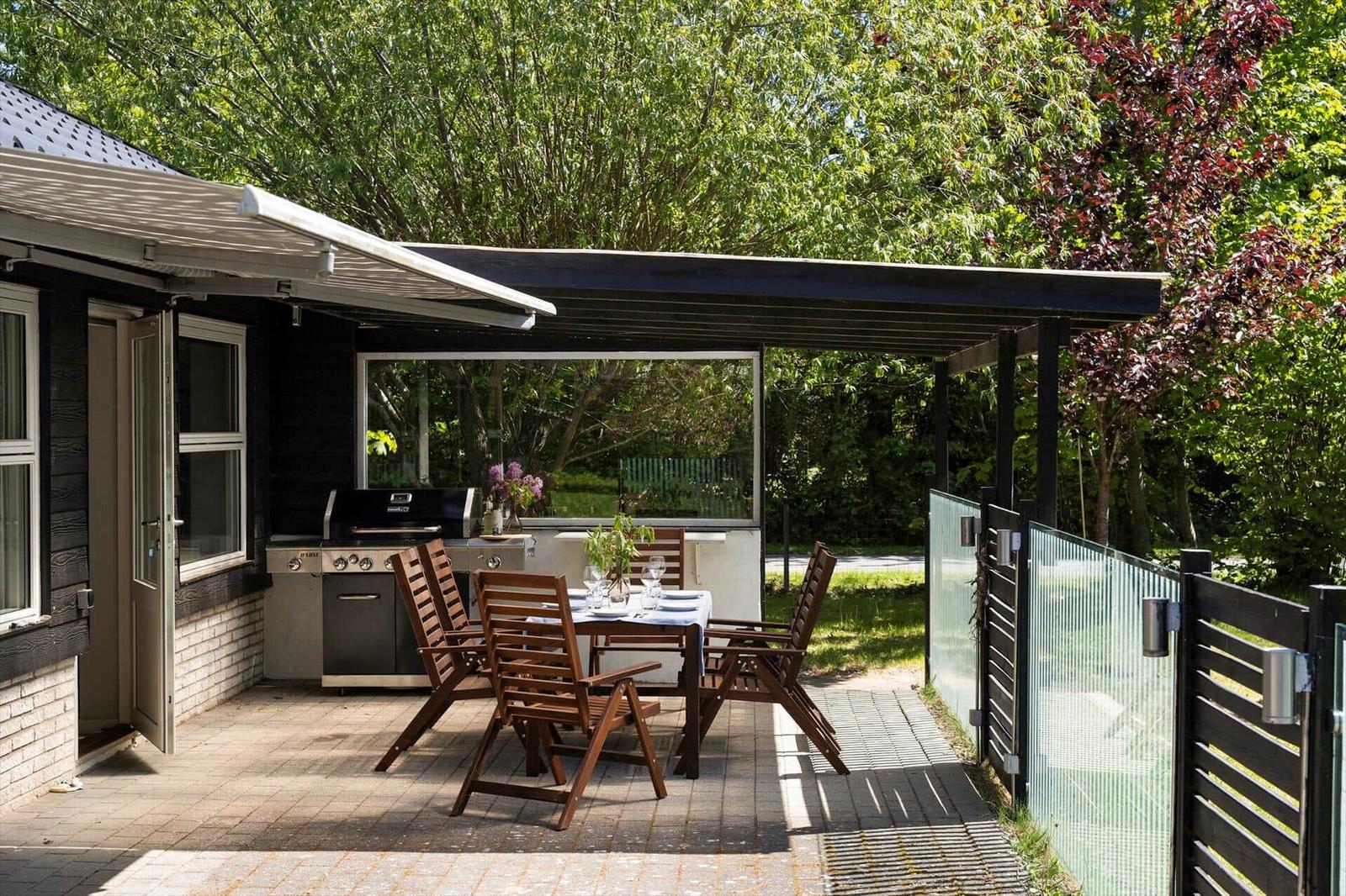 Terrace with grill, table and chairs under a pavilion. Surrounded by trees and a fence.