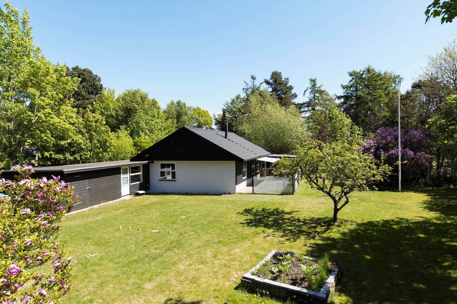 A house with a garden, surrounded by trees and shrubs. A flower bed and a tree are visible in the foreground.
