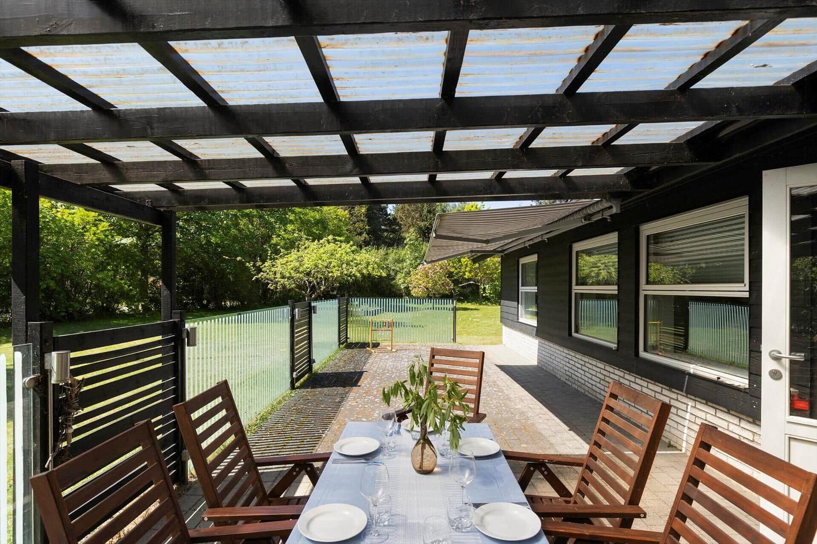 Terrace with table and chairs under a canopy. Background: greenery and house wall.