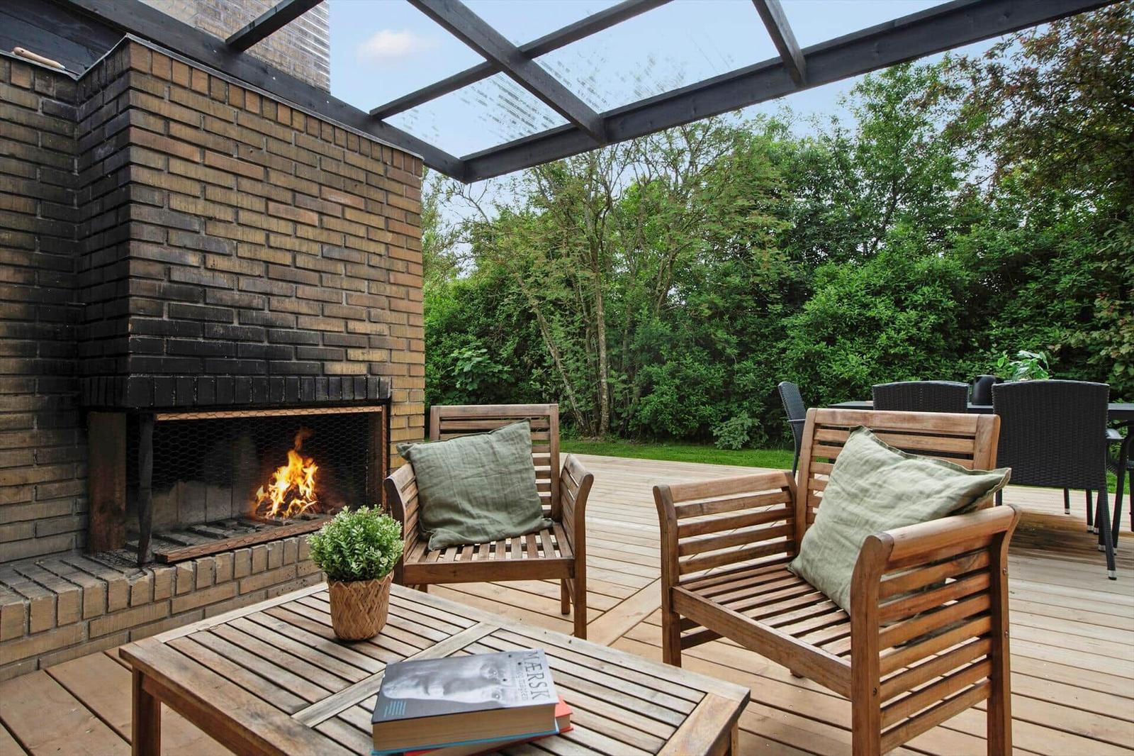 Outdoor area with wooden benches, fireplace, and table. Background: green trees.