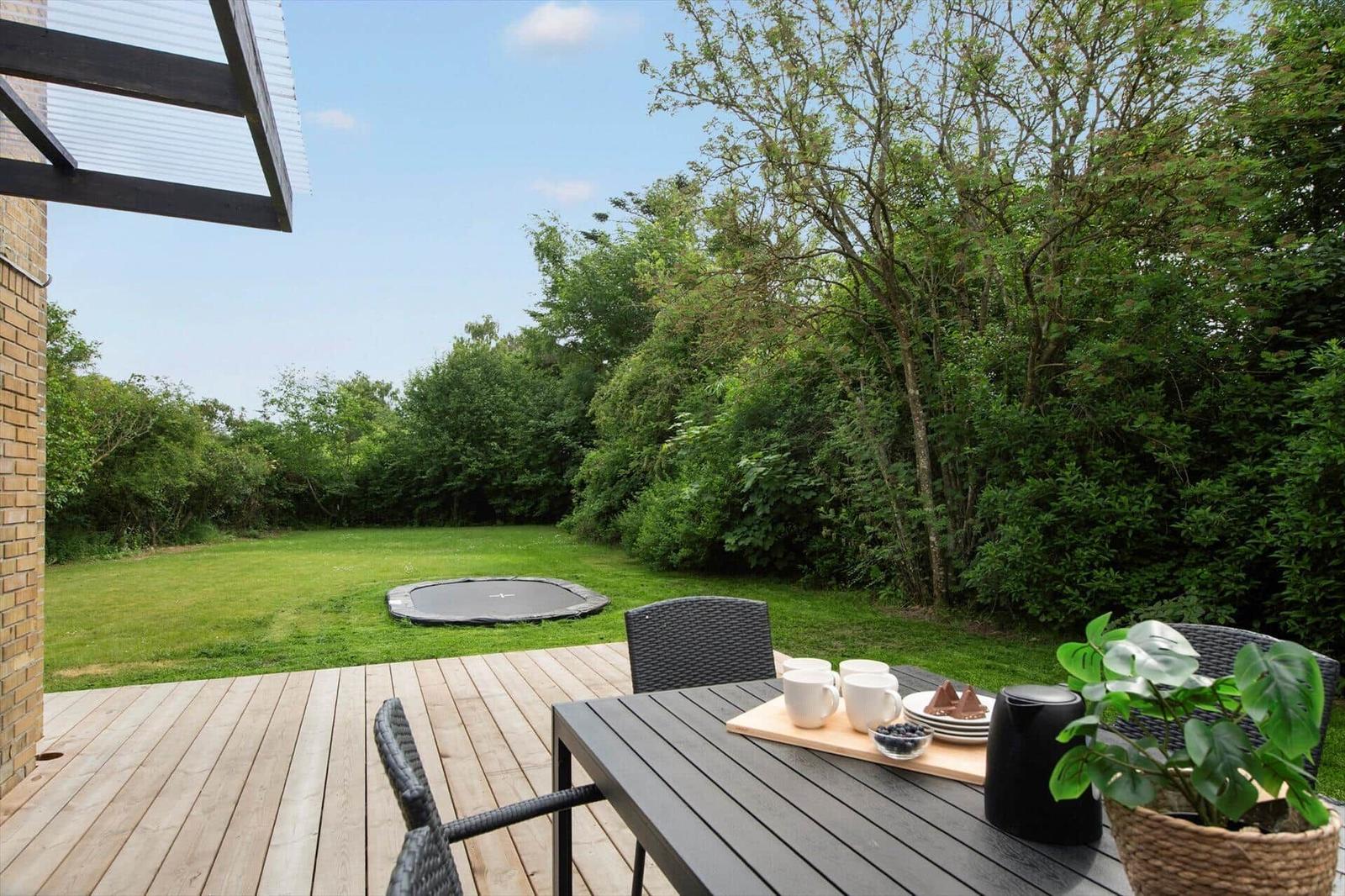 Deck with table, chairs, and plant. Lawn with trampoline and trees in background.