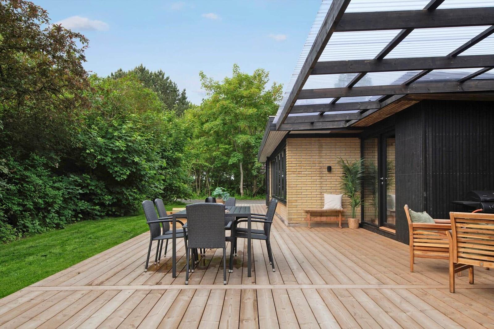 Wooden terrace with seating and grill. Background: green garden and house with glass roof.