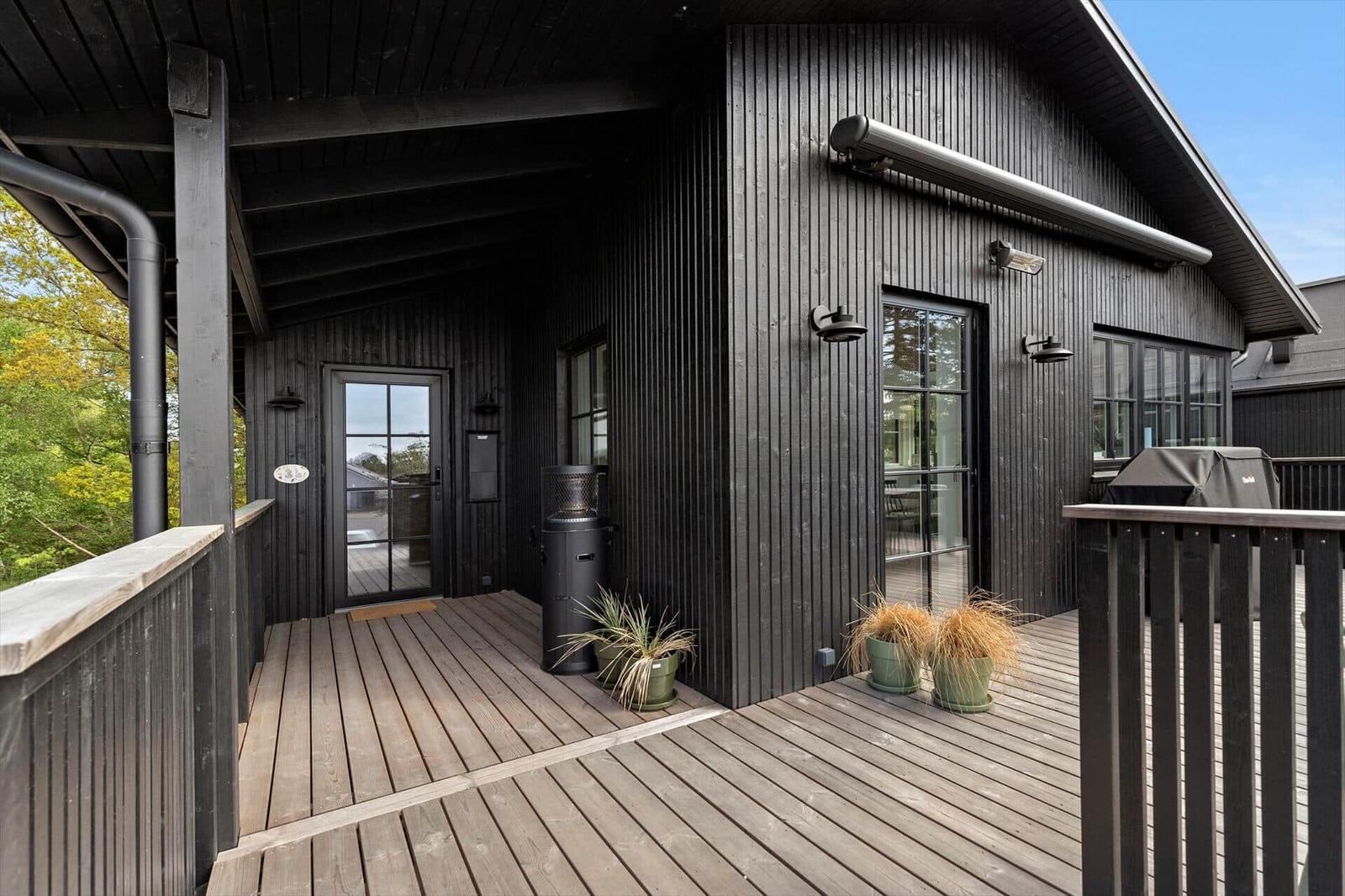 Black wooden house with terrace, grill, and plants. Entrance door and windows visible.