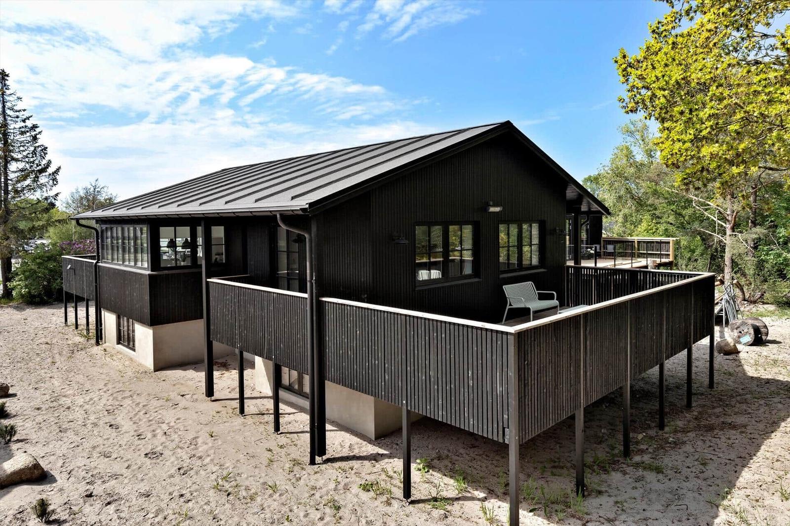 Black wooden house with balcony and windows, surrounded by trees and sand.