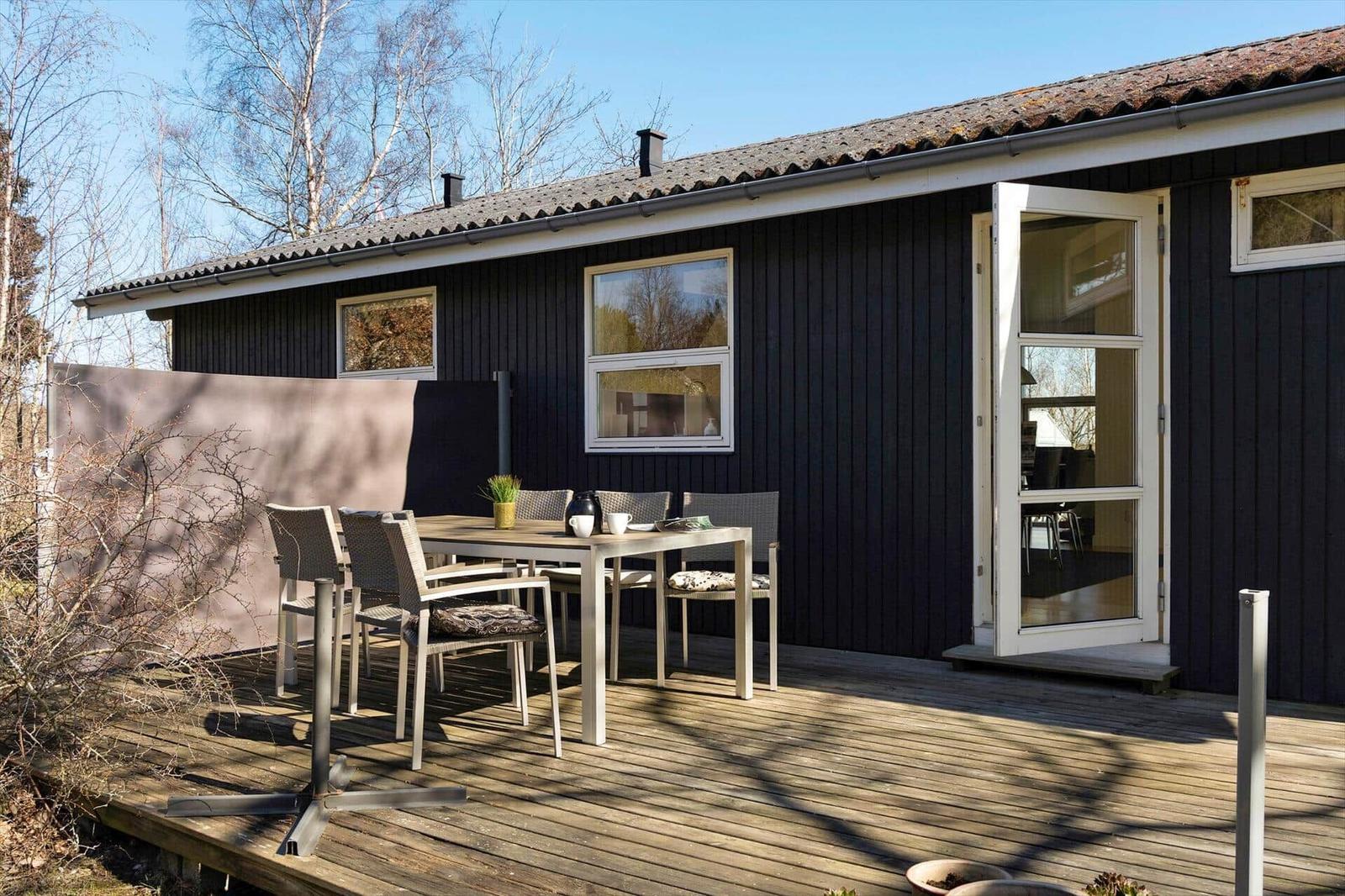 Holzterrasse mit Tisch und Stühlen vor einem dunklen Holzhaus mit offener Tür.