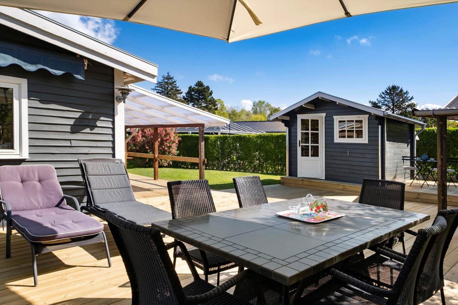 Deck with table, chairs, and umbrella. Small house and green garden in background.