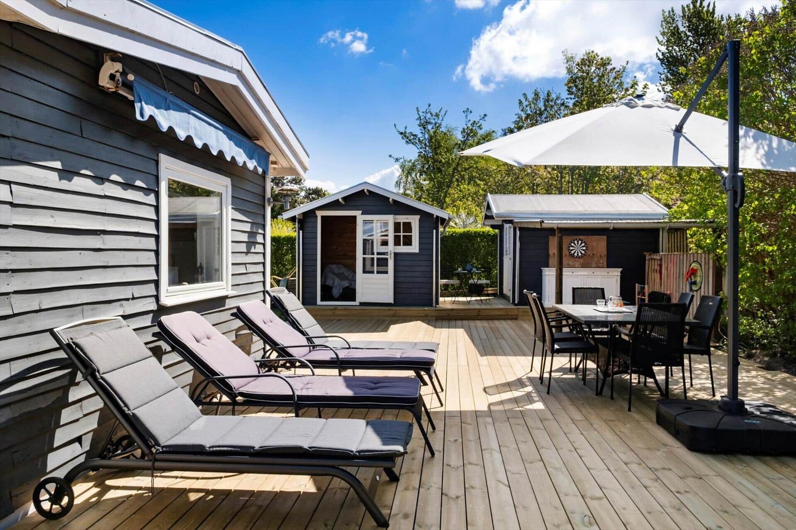 Deck with loungers, table, and umbrella. Background: wooden shed and grill area.