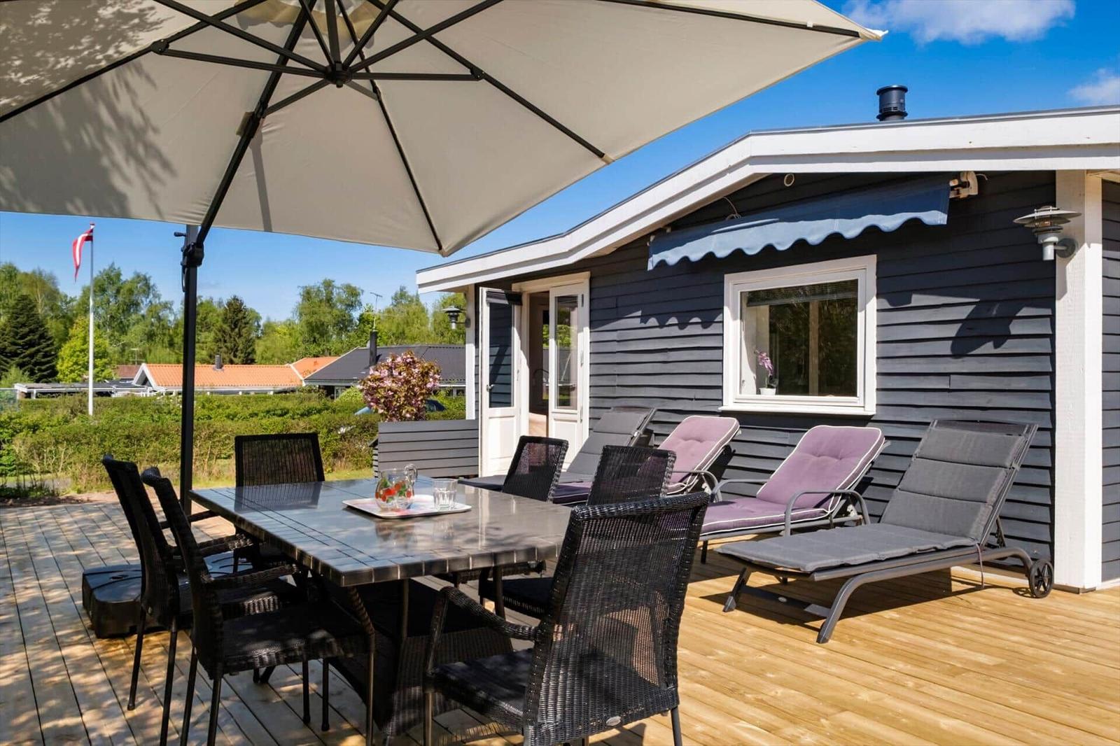 Deck with table, chairs, and umbrella. Background: house with window and lounge chairs.