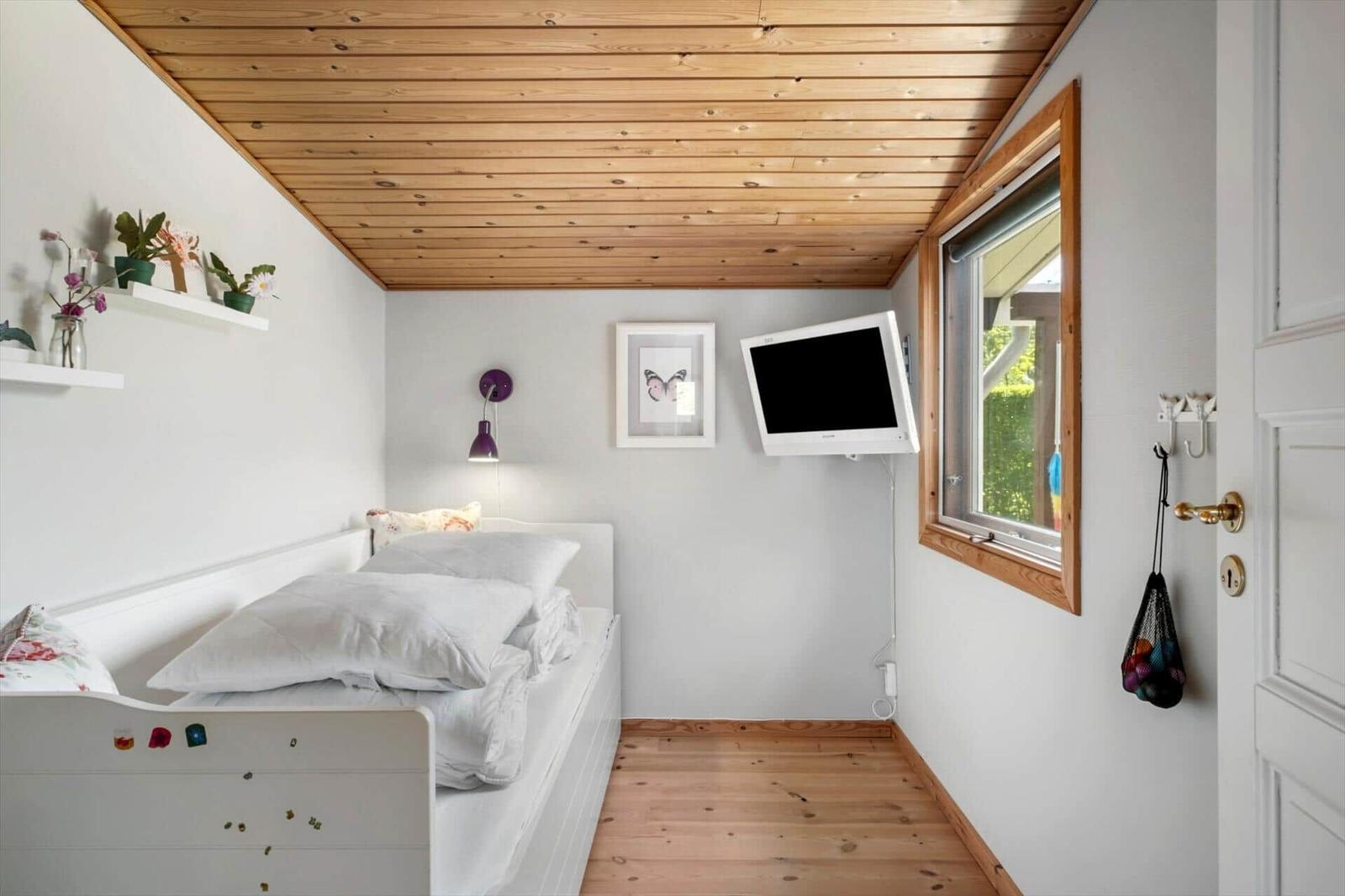 A bedroom with a white bed, wooden floor and ceiling. A TV is mounted on the wall.