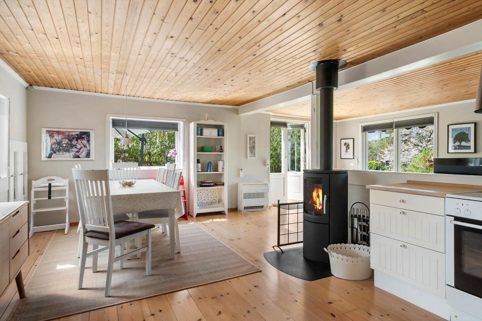 A room with dining area, wooden ceiling, fireplace, and kitchen counter. Windows show garden.