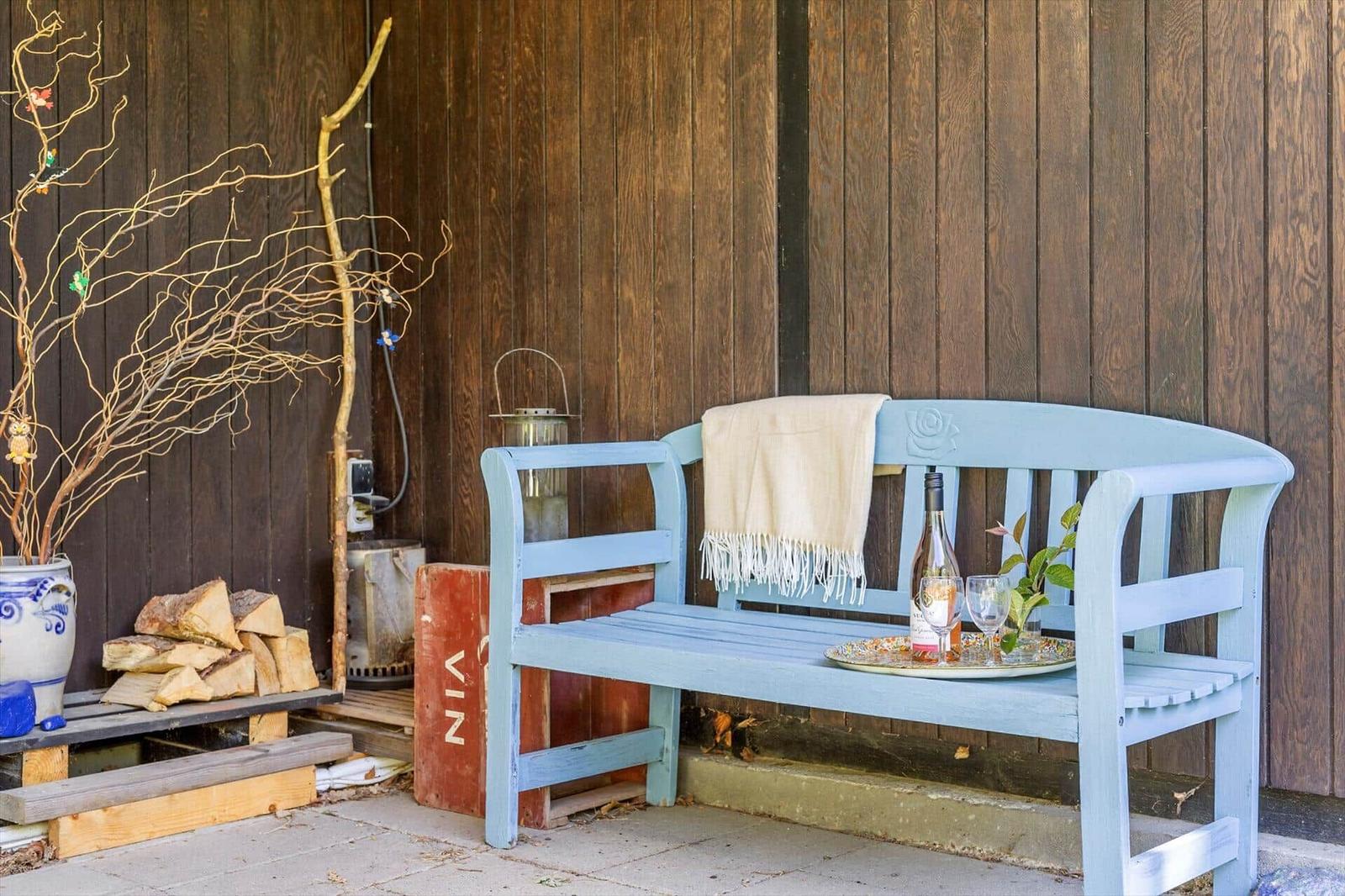 Blue wooden bench with blanket and drink against wood wall. Beside: woodpile and decorative branches.