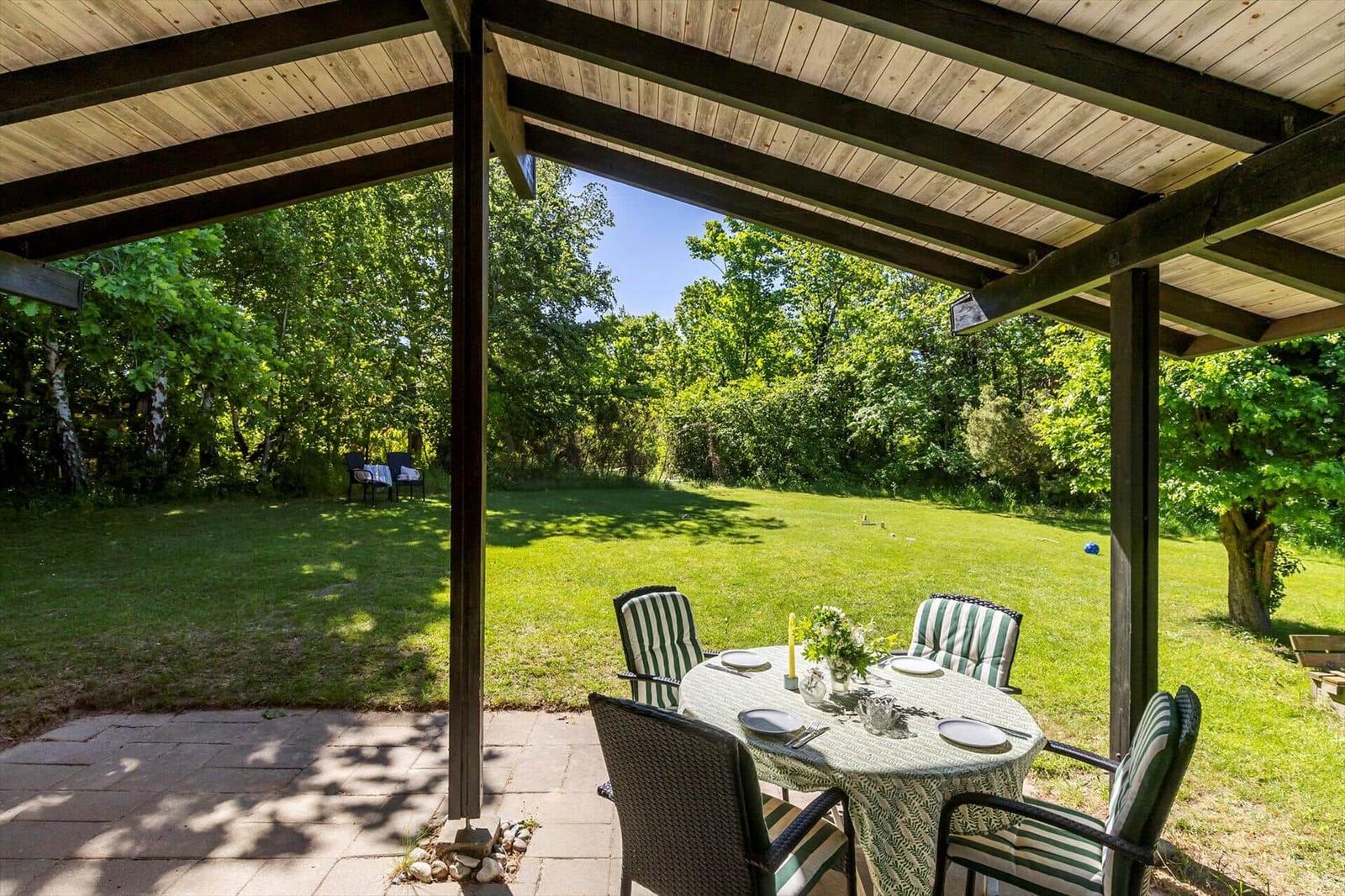 Covered outdoor area with table and chairs. Lawn and trees in the background.
