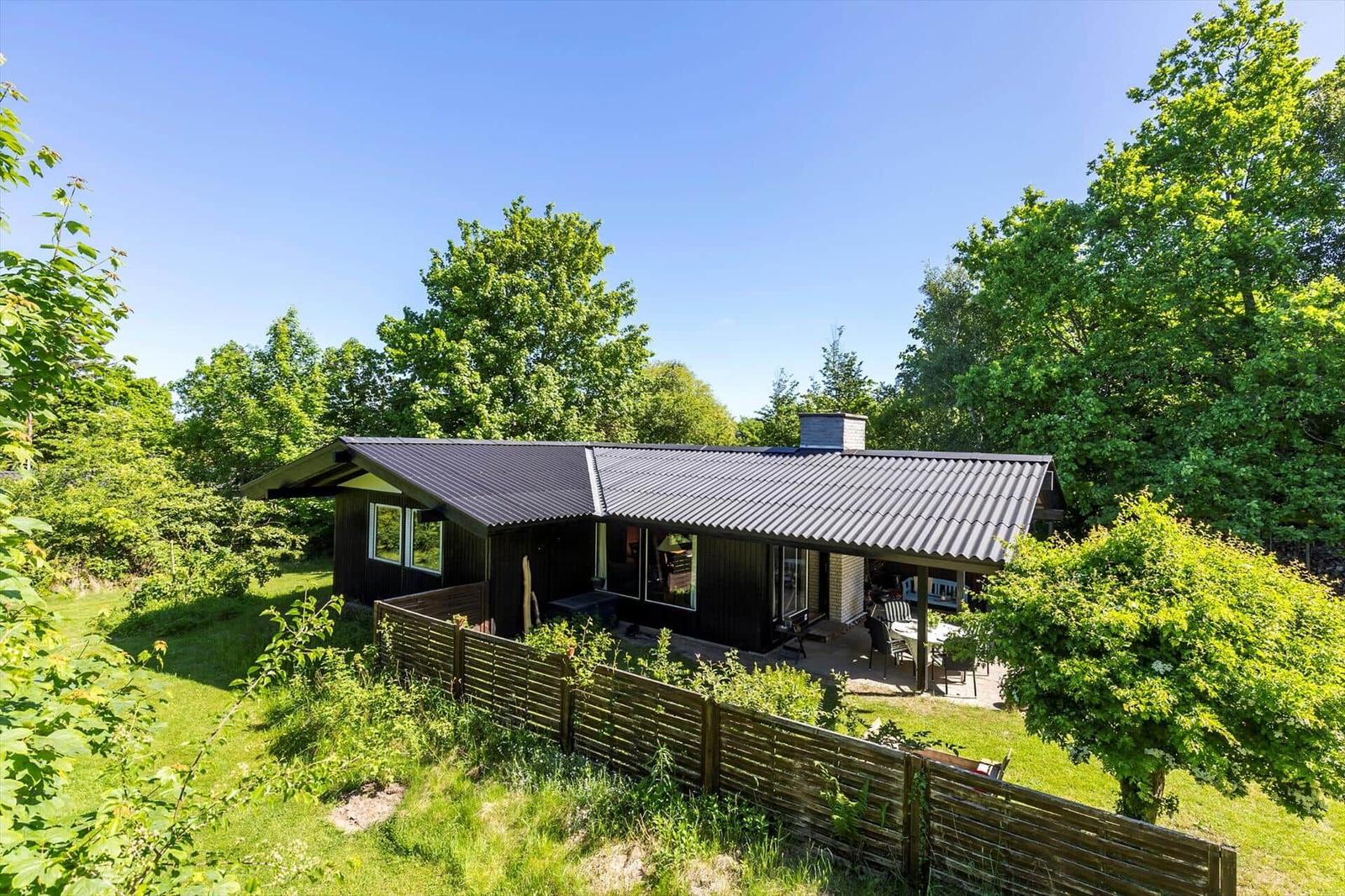 A house with dark cladding and roof, surrounded by greenery and trees. A terrace with table and chairs.
