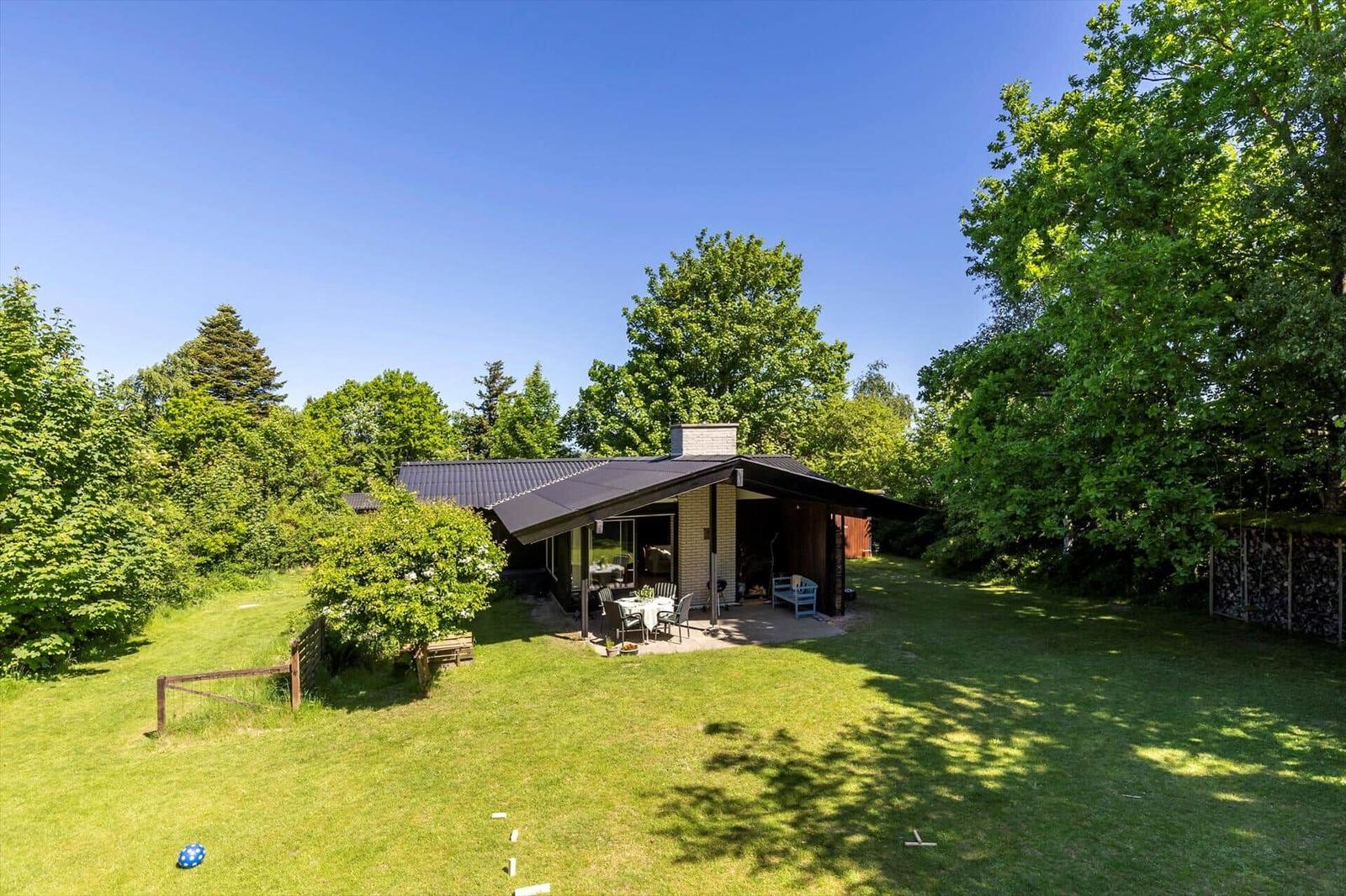 Modern villa with terrace, garden, and surrounding trees under blue sky.