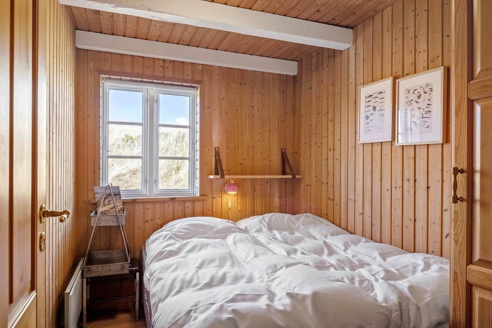 Bedroom with wooden walls, bed, and window. Two pictures hang on the wall.