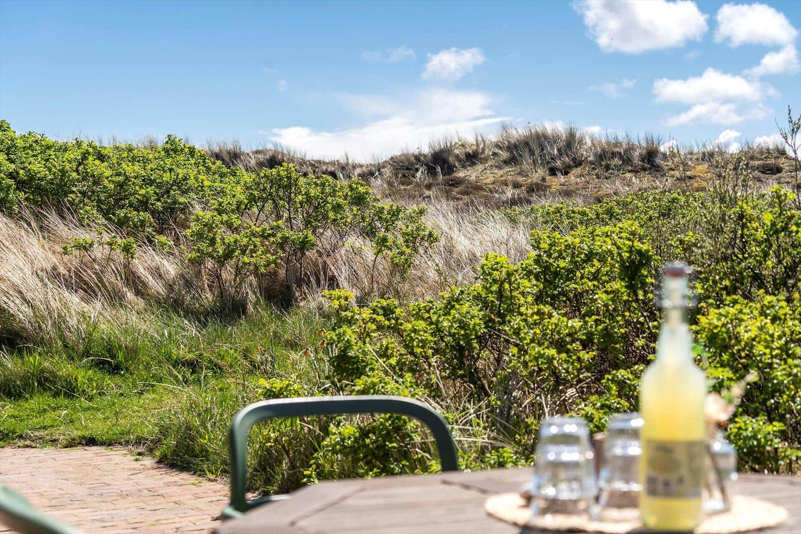 Outdoor area with table and chair, view of dunes and greenery under blue sky.