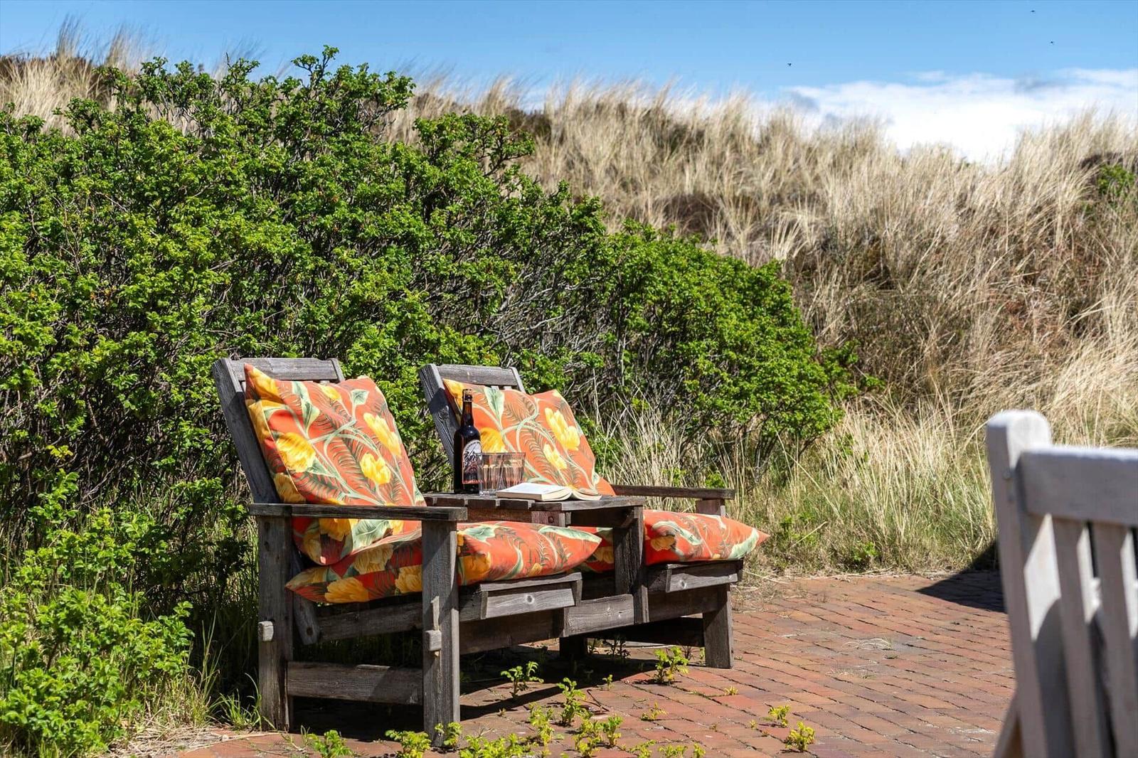 Two wooden chairs with colorful cushions stand on a brick path before grass and bushes.