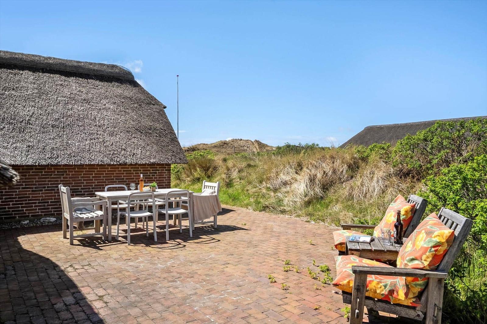 Terrace with table, chairs, and seating areas in front of a thatched roof house.