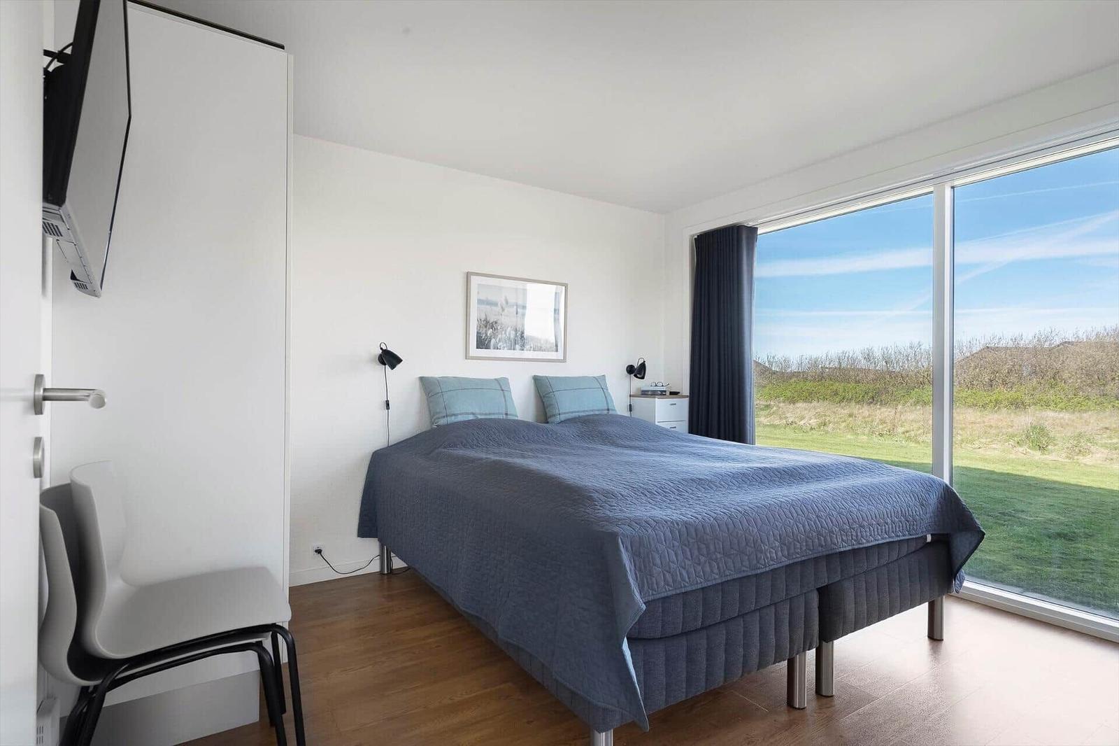 Schlafzimmer mit Bett, TV und großem Fenster mit Blick auf Grünflächen.
