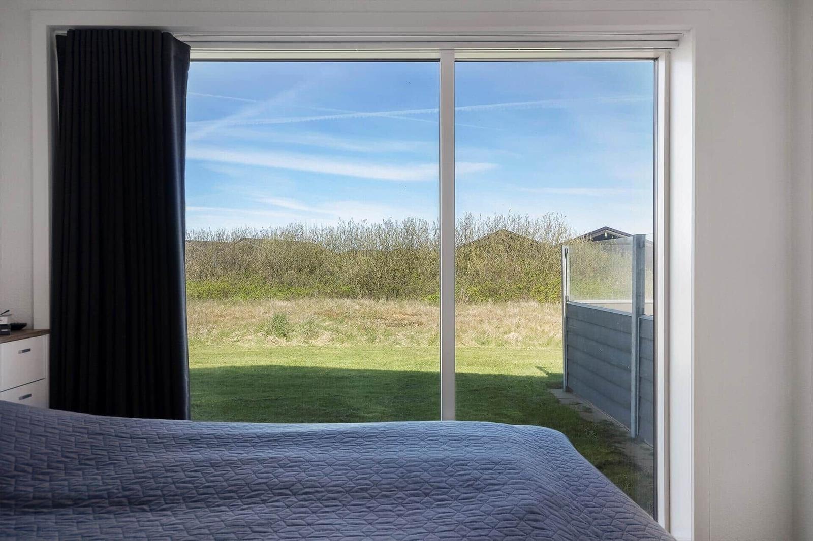 Schlafzimmer mit Blick auf grünen Garten und Himmel durch große Fenster.